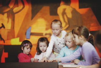 Zdjęcie. Grupa dzieci w skupieniu pracuje nad zadaniem. W tle fragment ściennego obrazu Fangora w Muzeum Warszawy.