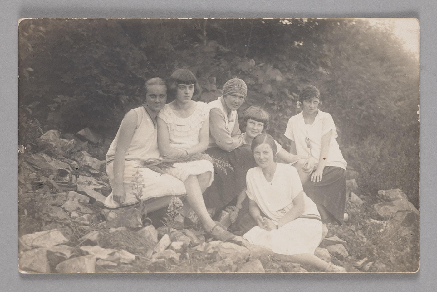 Zdjęcie grupy sześciu kobiet w białych sukienkach i koszulach, siedzących na zewnątrz na tle zieleni.