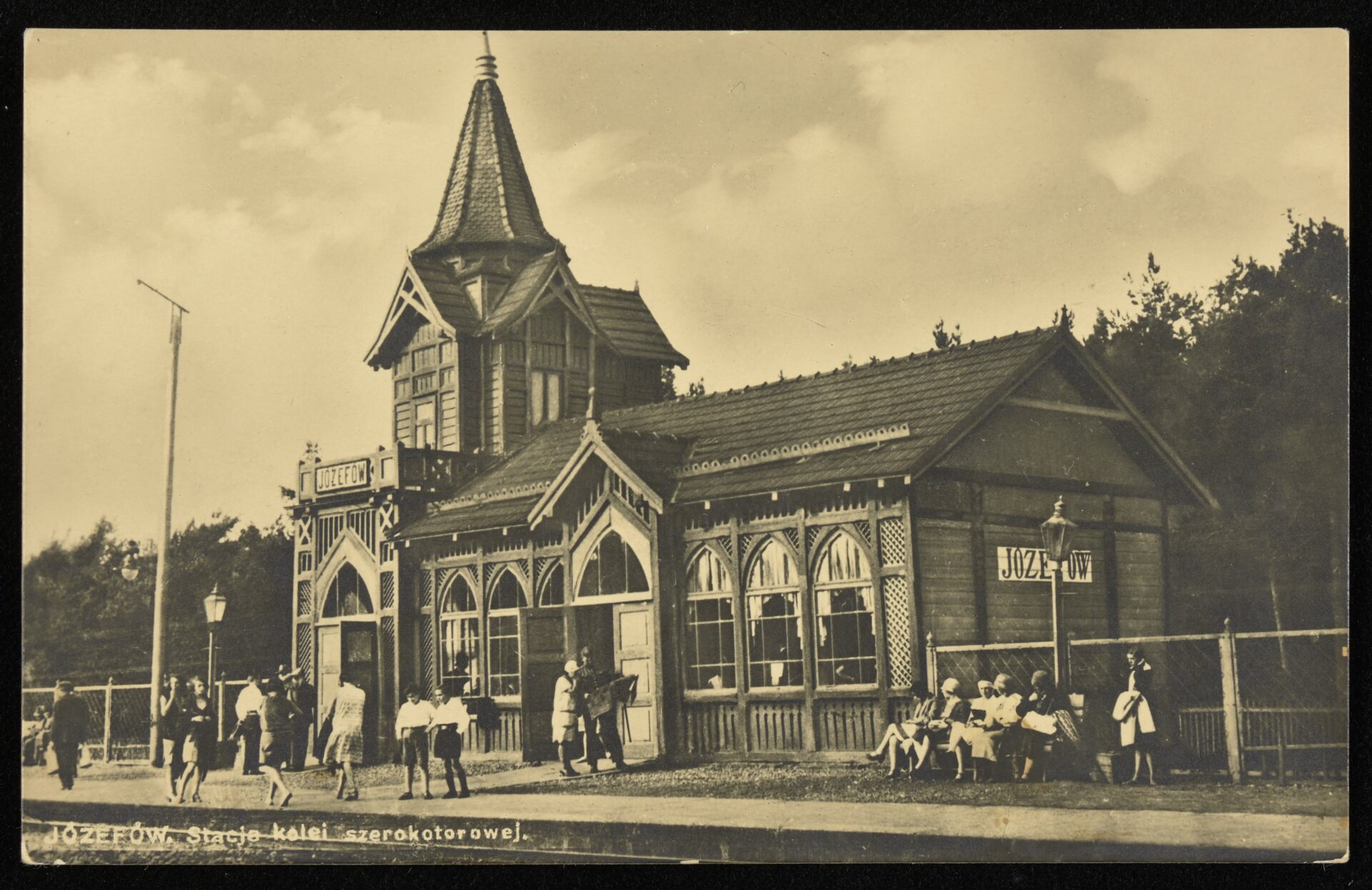 Zdjęcie archiwalne, czarno-białe. Drewniany budynek stacji kolejowej w Józefowie. Przed nim zgromadzeni ludzie.