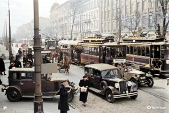 Fotografia kolorowa ulica Warszawy. Tramwaje, samochody, dorożki, spacerujący ludzie.