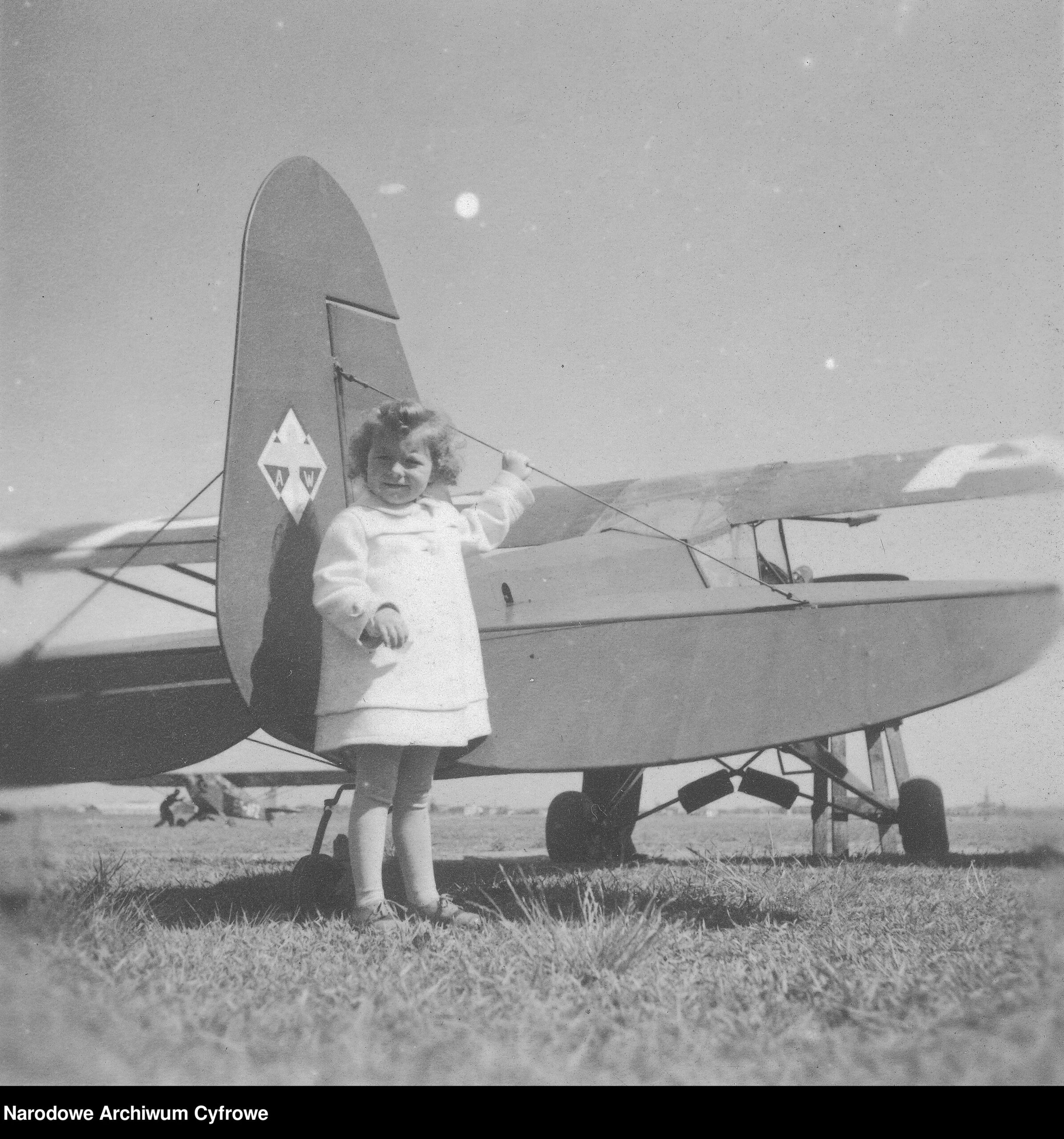 Czarno białe zdjęcie. Mała dziewczynka ubrana na biało stoi przy starym samolocie.