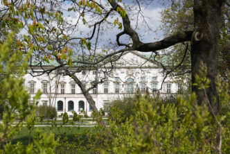 Zdjęcie. Zza drzew wyłania się biała fasada Pałacu Krasińskich.