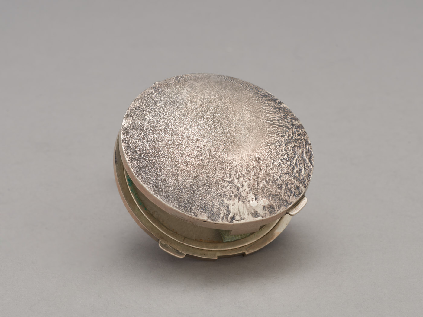 Zdjęcie srebrnej, okrągłej puderniczki. Na wieku nieregularna faktura, powstała poprzez rozgrzanie i stopienie wierzchniej warstwy metalu.