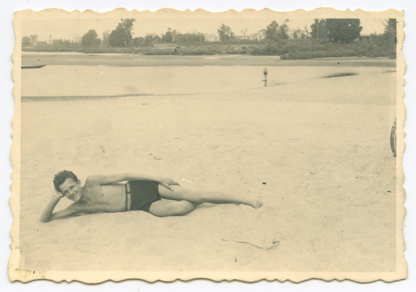 Fotografia. Nad brzegiem Wisły, na piaszczystej plaży młody chłopak leży na prawym boku. Podpiera głowę na ręce, uśmiecha się. Ubrany tylko w ciemne krótkie spodenki. W tle niewysokie drzewa i krzewy.