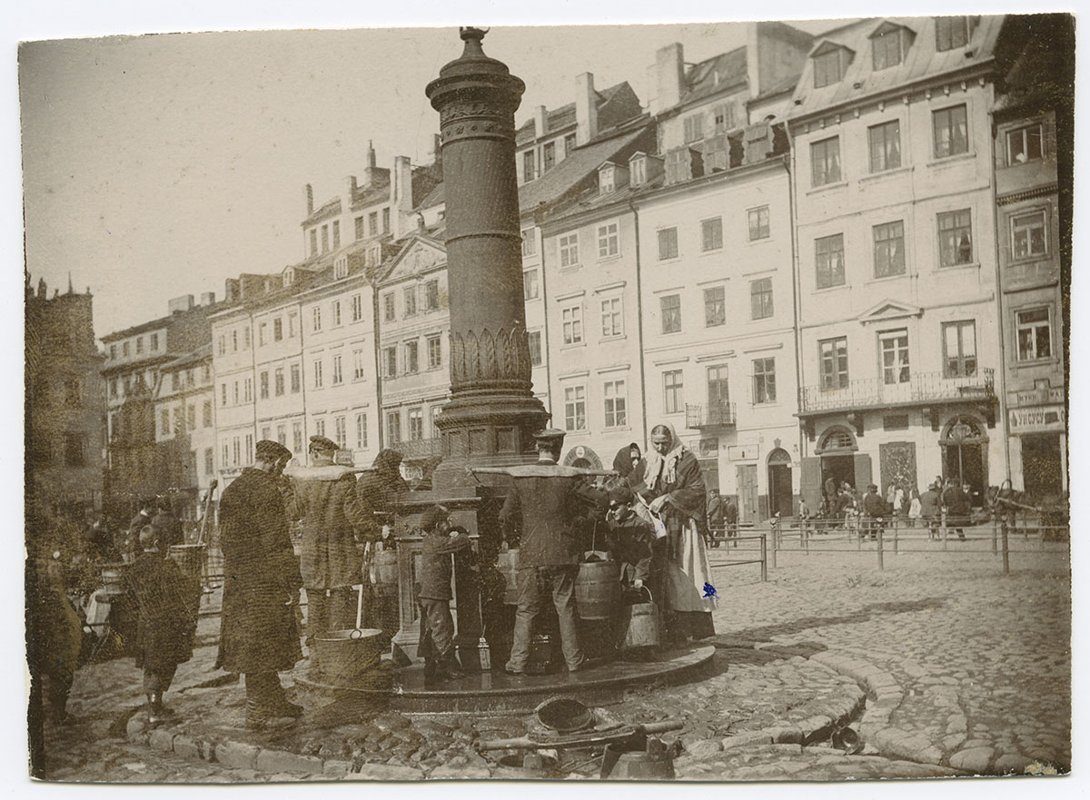 Czarno biała fotografia. Na pierwszym planie wysoka pompa przy Rynku Starego Miasta. Wokół niej mężczyźni, kobiety i dzieci napełniają wiadra wodą. W tle rząd kamienic.