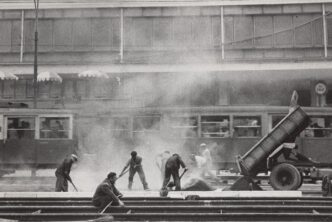 poziome czarno białe zdjęcie Edwarda Hartwiga, robotnicy kładą asfalt, rozprowadzają go łopatami, ciężarówka przechylona po prawej, nad nimi unosi się obłok