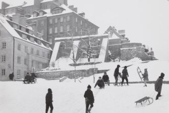 czarno białe zdjęcie, skarpa przy starym mieście, po lewej kamienice i fragmenty barbakanu, po prawej dzieci z sankami, wszystko pokryte śniegiem
