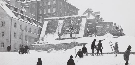 czarno białe zdjęcie, skarpa przy starym mieście, po lewej kamienice i fragmenty barbakanu, po prawej dzieci z sankami, wszystko pokryte śniegiem
