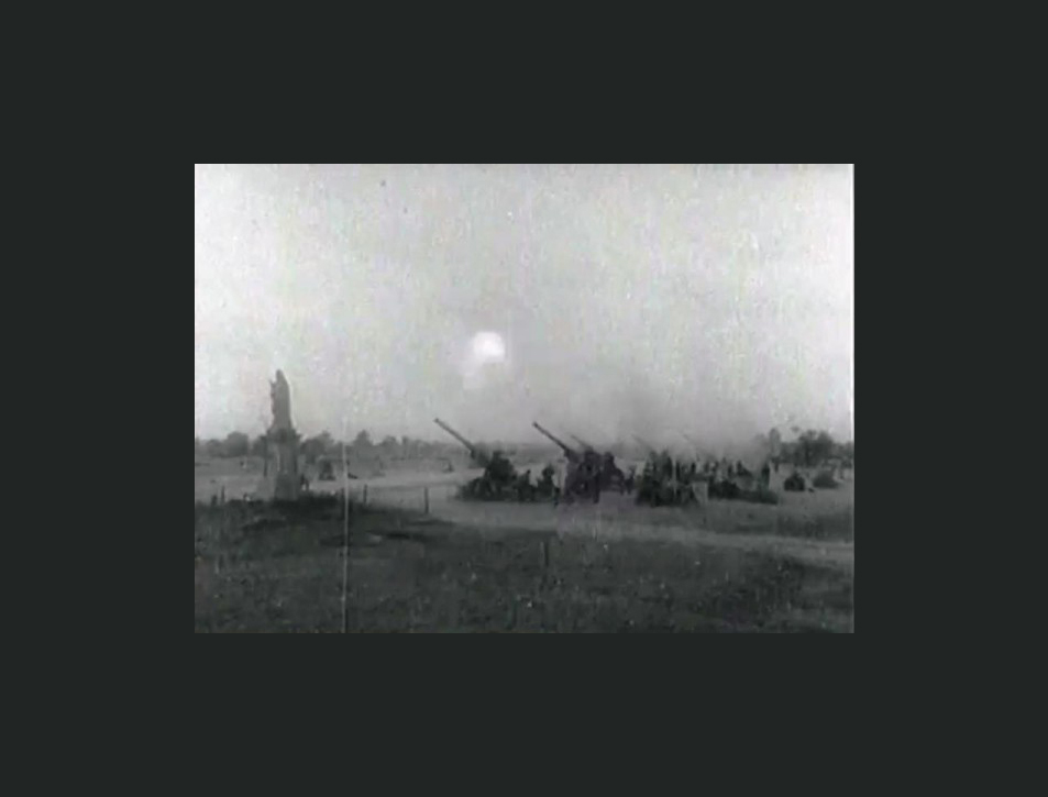 Czarno-białe zdjęcie w ciemnej ramie. W lewym rogu figura maryjna. Obok cztery działa w momencie oddawania wystrzału.