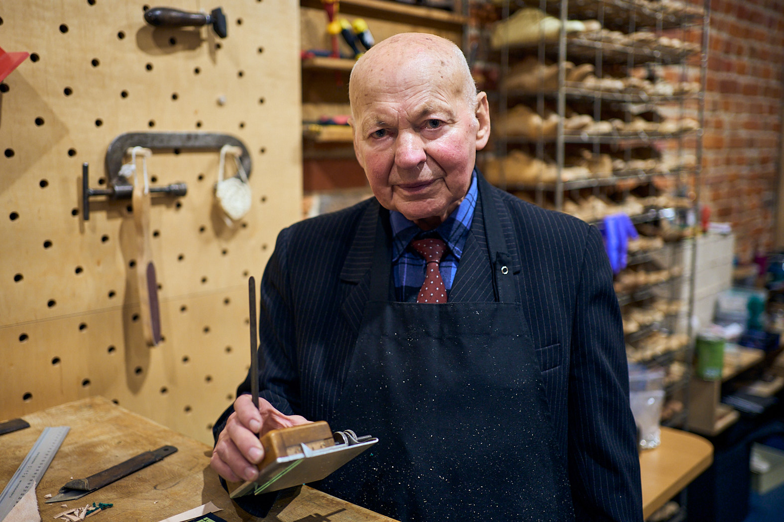 Zdjęcie. Starszy mężczyzna w garniturze w prążki i skórzanym fartuchu, trzymający narzędzie kaletnicze w dłoni.