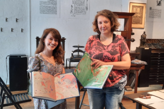 Na zdjęciu dwie kobiety prezentują kolorowe szkicowniki, uśmiechają się. W tle, po prawej stronie drewniana prasa drukarska.
