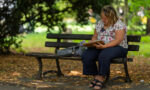 Zdjęcie. Kobieta siedzi w cieniu na ławce w parku i rysuje w szkicowniku, który trzyma na kolanach.
