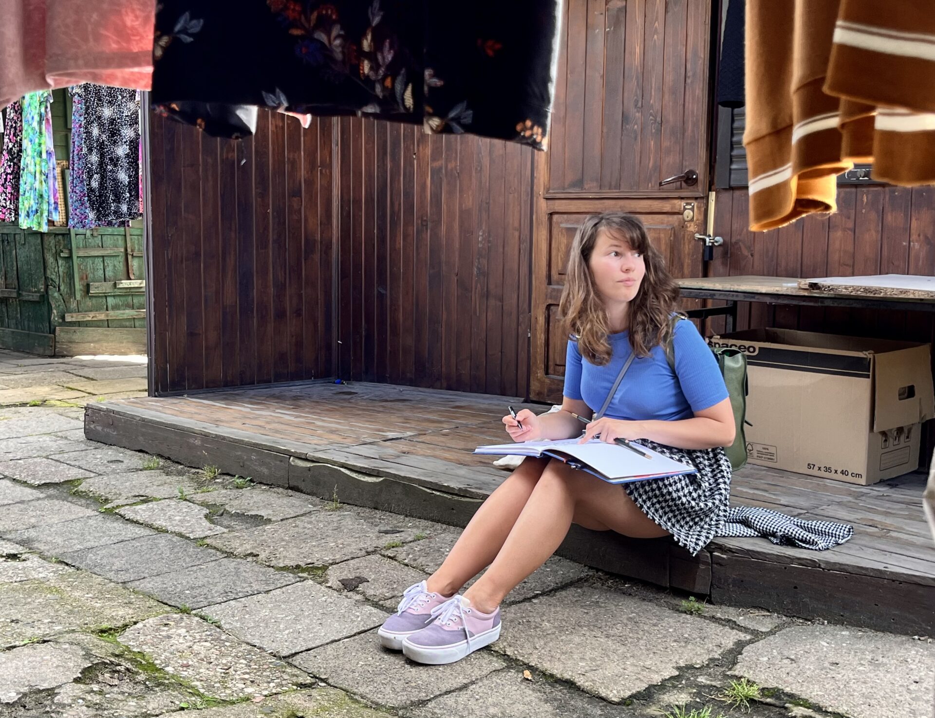 Zdjęcie. Dziewczyna w niebieskim tiszercie, spódnicy w kratkę i sportowych butach siedzi na drewnianym podeście przed sklepem budką na Bazarze Różyckiego. Na kolanach trzyma notatnik. Patrzy w bok.