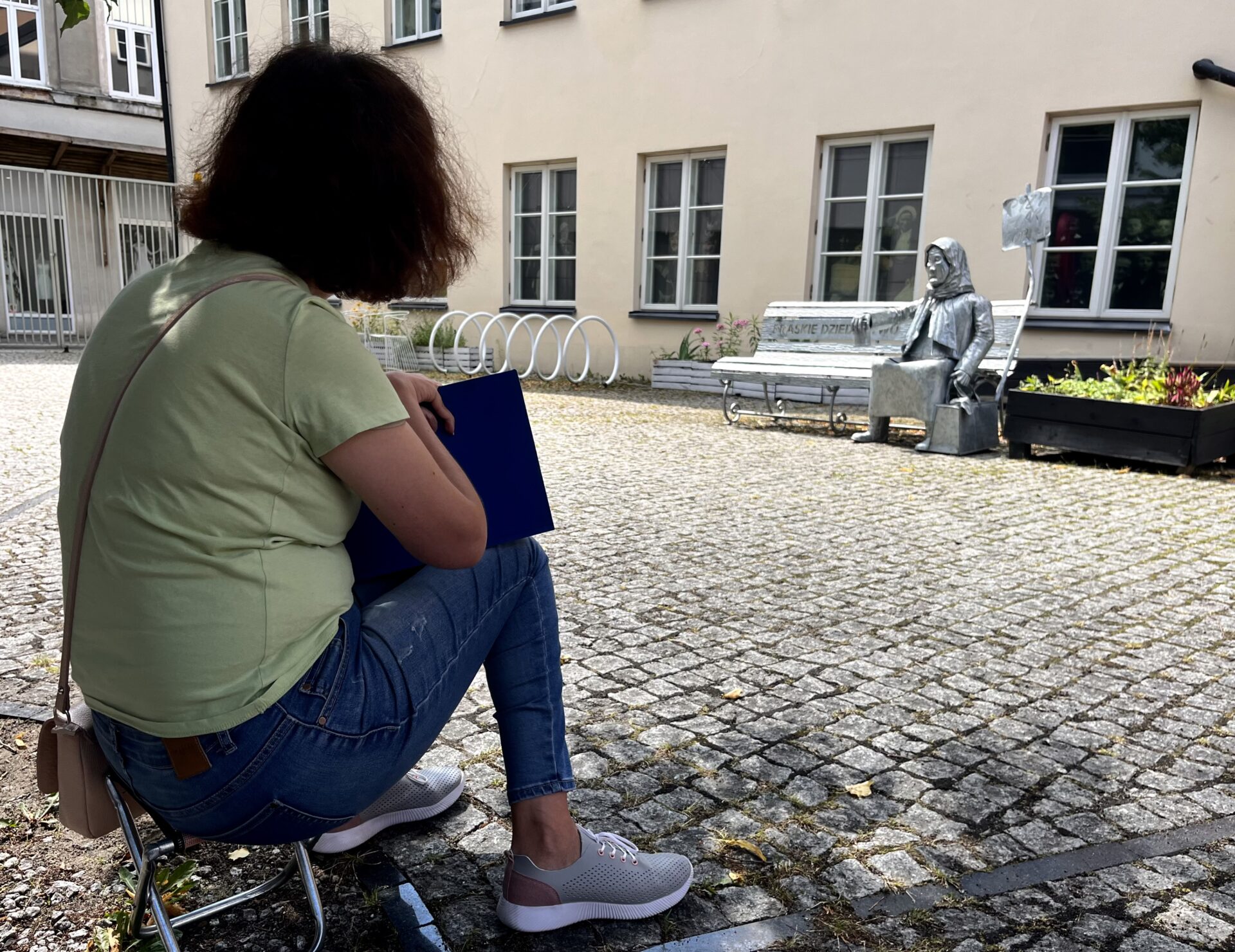 Zdjęcie. Dziewczyna kuca w cieniu na dziedzińcu Muzeum Warszawskiej Pragi i patrzy na ławkę rzeźbę starszej kobiety w chuście.
