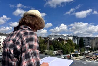 Zdjęcie. Mężczyzna w dłuższych włosach, kapeluszu i koszuli w kratę stoi na tarasie Muzeum Warszawskiej Pragi z widokiem na Bazar Różyckiego i rysuje. Słoneczne, niebieskie niebo z chmurami.