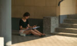 Zdjęcie. Dziewczyna siedzi w cieniu przy schodach do budynku. Na kolanach trzyma szkicownik, rysuje.