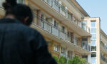 Zdjęcie. Na pierwszym planie nieostra pochylona postać mężczyzny stojącego tyłem. W tle wyraźna kamienica mieszkalna z balustradami.