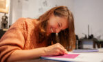 Zdjęcie. Roześmiana dziewczyna rysuje coś na kartce leżącej przed nią na blacie stołu.